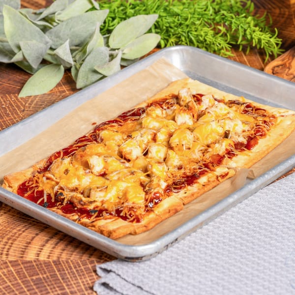 OSO's BBQ chicken flatbread pizza, topped with melted cheese, on a baking sheet.