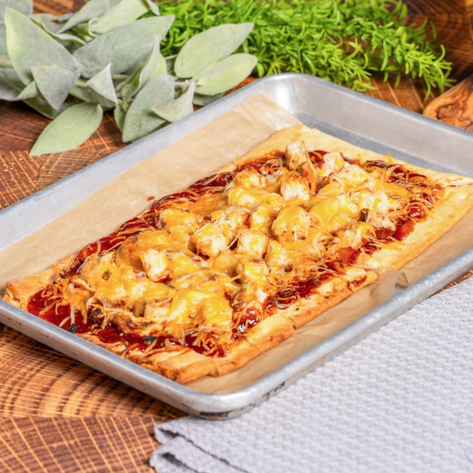 OSO's BBQ chicken flatbread pizza, topped with melted cheese, on a baking sheet.