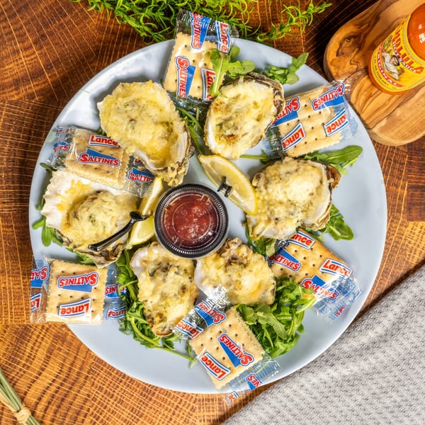 A plate of baked oysters topped with cheese, served with crackers, lemon, and cocktail sauce.