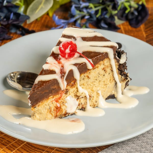 A slice of chocolate and peanut butter cheesecake, topped with white drizzle and a maraschino cherry.