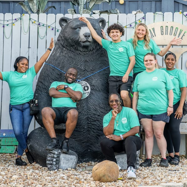 Seven OSO staff members in teal shirts posing with a large black bear statue outdoors.