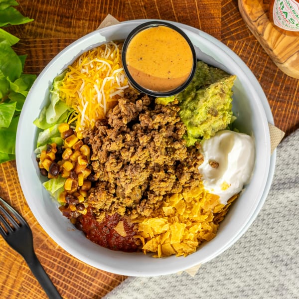 Taco salad bowl with ground beef, cheese, guacamole, sour cream, salsa, corn, and tortilla chips.