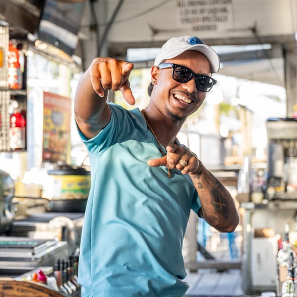 Smiling man in sunglasses and a light blue shirt pointing at the viewer in a restaurant setting.