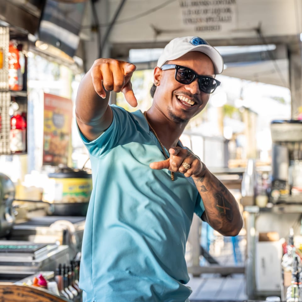 Smiling man in sunglasses and a light blue shirt pointing at the viewer in a restaurant setting.
